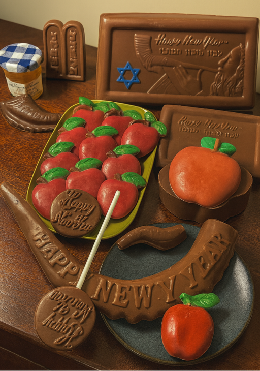 Rosh Hashana chocolate collection displayed on a wooden buffet table, featuring handcrafted chocolate shofars, red chocolate apples with green leaves, apple lollipops, ‘Happy New Year’ chocolate plaques, and a mini Bonne Maman honey jar. The chocolates show natural textures and imperfections, styled like a luxury magazine spread with warm, natural lighting
