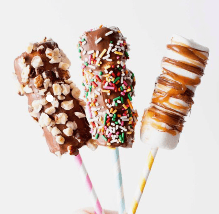 Three Chocolate Covered Marshmallow Pops on striped sticks, topped with nuts, caramel, and rainbow sprinkles for Purim celebrations.