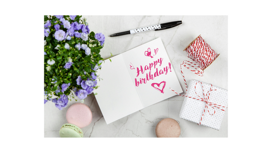 Happy birthday greeting card on a marble table with pastel macarons, purple flowers, a wrapped gift box, and festive twine — perfect birthday celebration scene for unique chocolate gift ideas