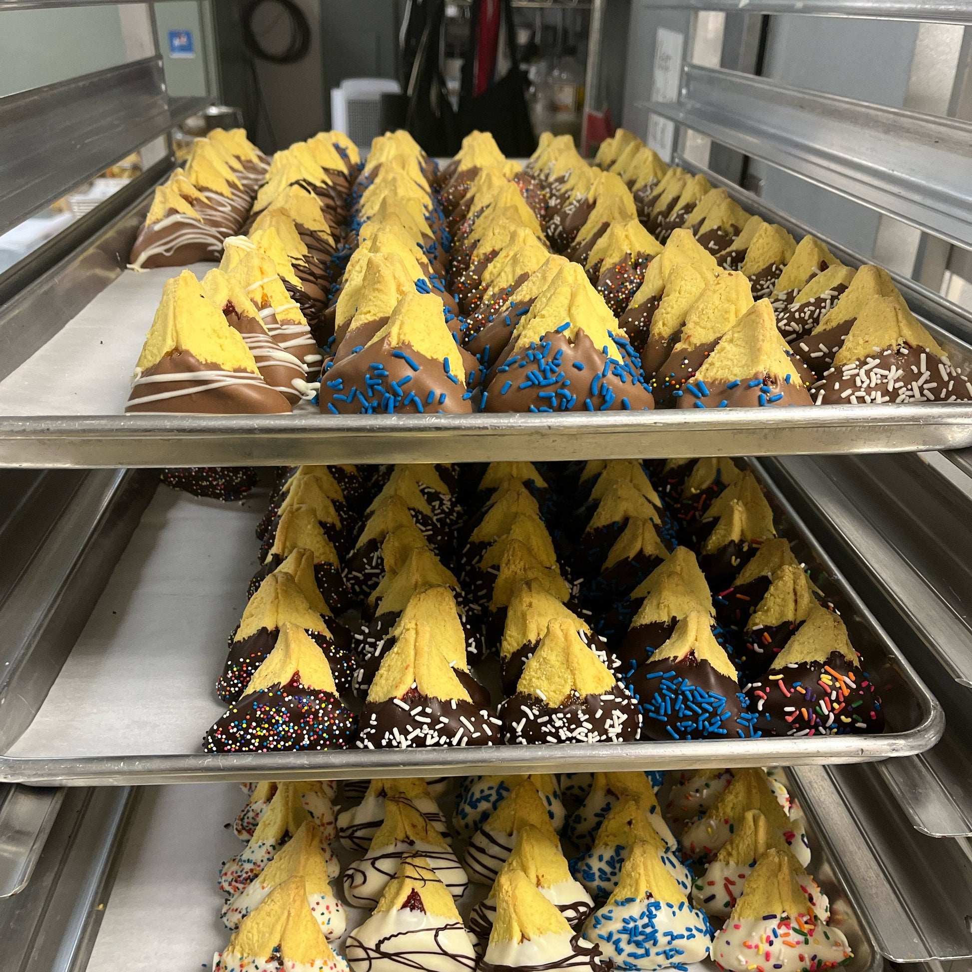 Assorted chocolate-dipped hamantaschen for Purim in Miami with colorful sprinkles on bakery trays for Small Purim Basket gift. Perfect for festive celebrations.