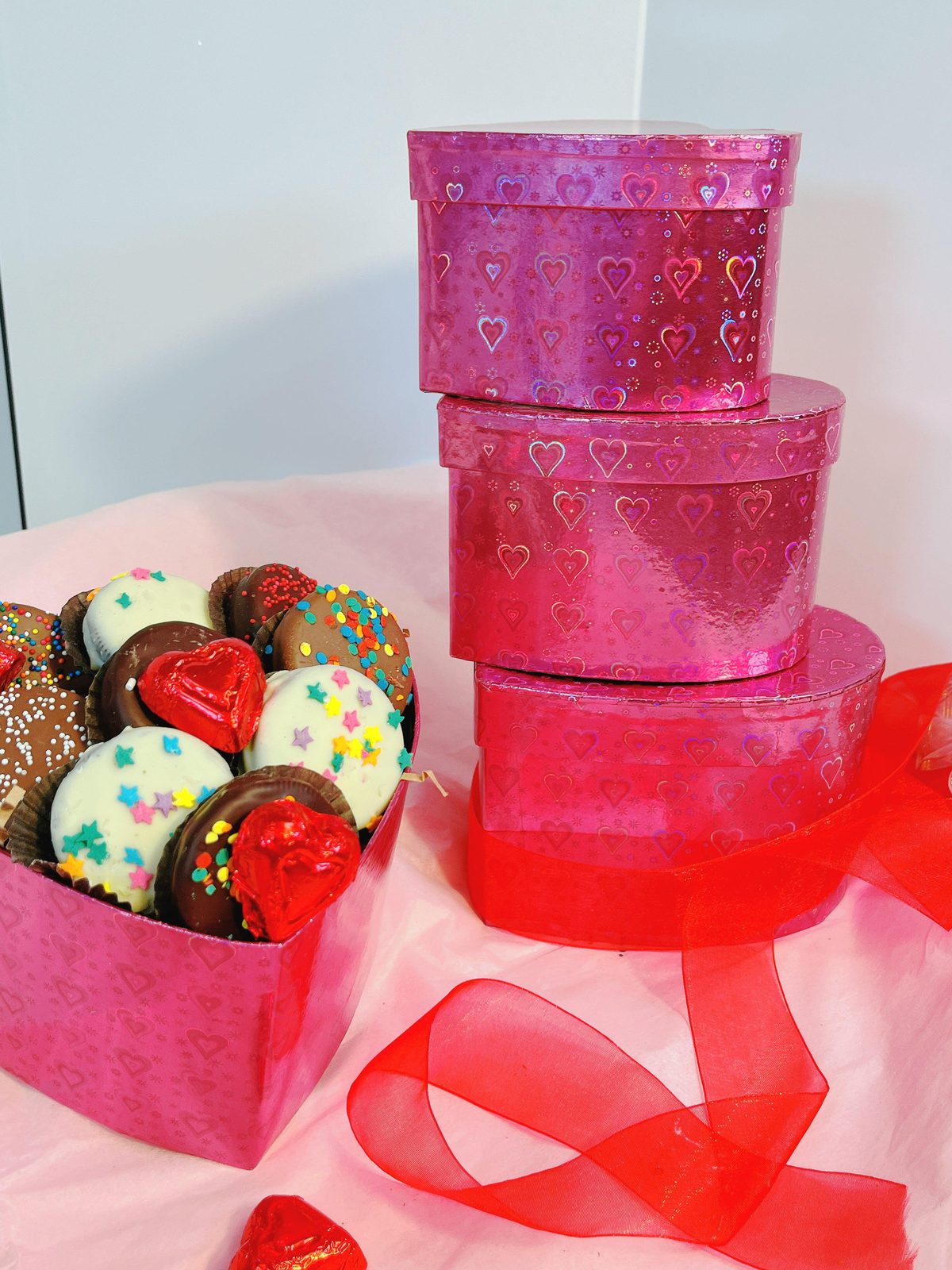 Stacked 3-tier heart-shaped gift boxes with glossy pink hearts design, showcasing elegant chocolate and truffle assortment beside them.