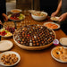 Assorted  chocolate and pastries in a large basket on a table with various other food items and people around.