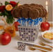 Silver mosaic gift box filled with about 18 chocolate lollipops embossed with 'Happy New Year,' each wrapped in clear cello with blue ribbon curls. The box is set on an elegant Rosh Hashanah holiday table with apples, pomegranates, honey jar, gold cutlery, roses, and a lit candle in the background.