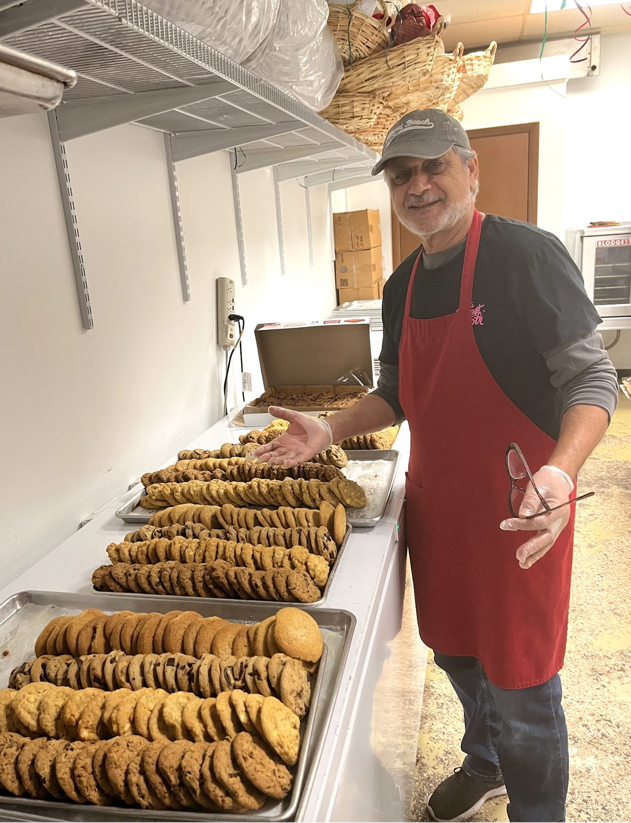 Zalman, expert cookie connoisseur in a red apron and a "key west" t-shirt and  cap, standing in the kitchen at The Sweet Tooth next to cooling racks filled with dozens of freshly baked cookies. 