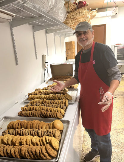 Zalman, expert cookie connoisseur in a red apron and a "key west" t-shirt and  cap, standing in the kitchen at The Sweet Tooth next to cooling racks filled with dozens of freshly baked cookies. 