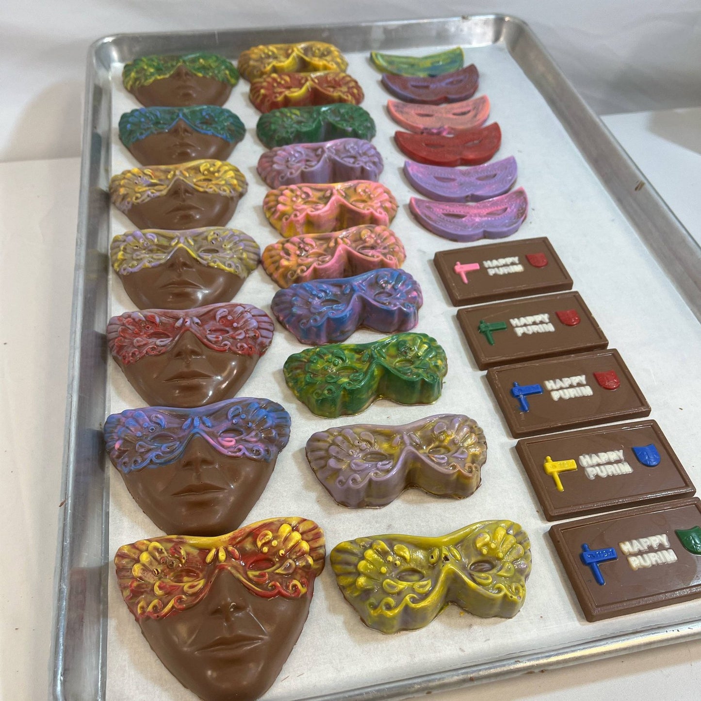 Tray of colorful hand-painted Chocolate Purim Masks and themed chocolate bars with 'Happy Purim' text, ideal for festive candy assortments.